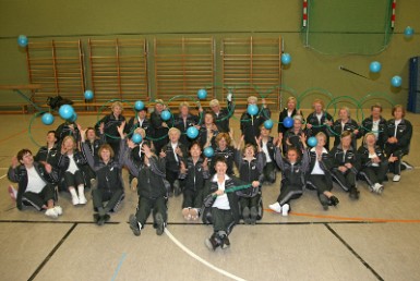 Gruppenbild der Gymnastik-Damen sitzend mit Dehnbändern, Reifen und in die Luft geworfenen Bällen